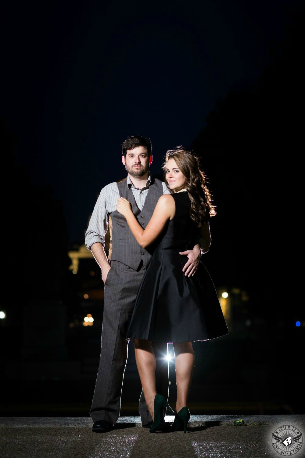 brown haired girl in black dress and black pumps smirks at camera while holding on to vest of brown haired guy wearing grey and white striped vest and pants at top of steps in darkness in this engagement photo at UT austin